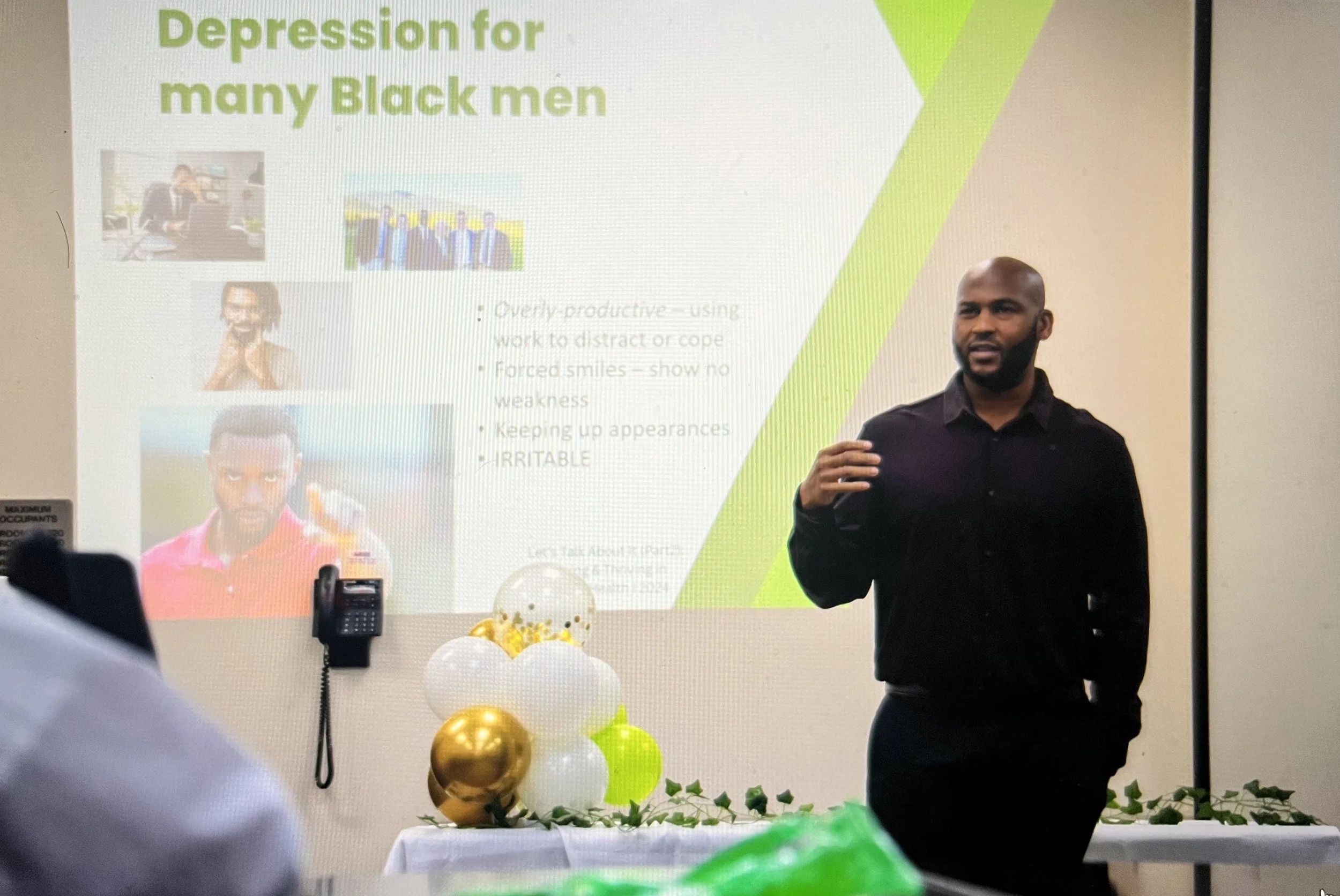 A man giving a presentation in a room decorated with gold, white, and green balloons. The projection behind him shows a slide titled "Depression for many Black men" with text and images of black men.