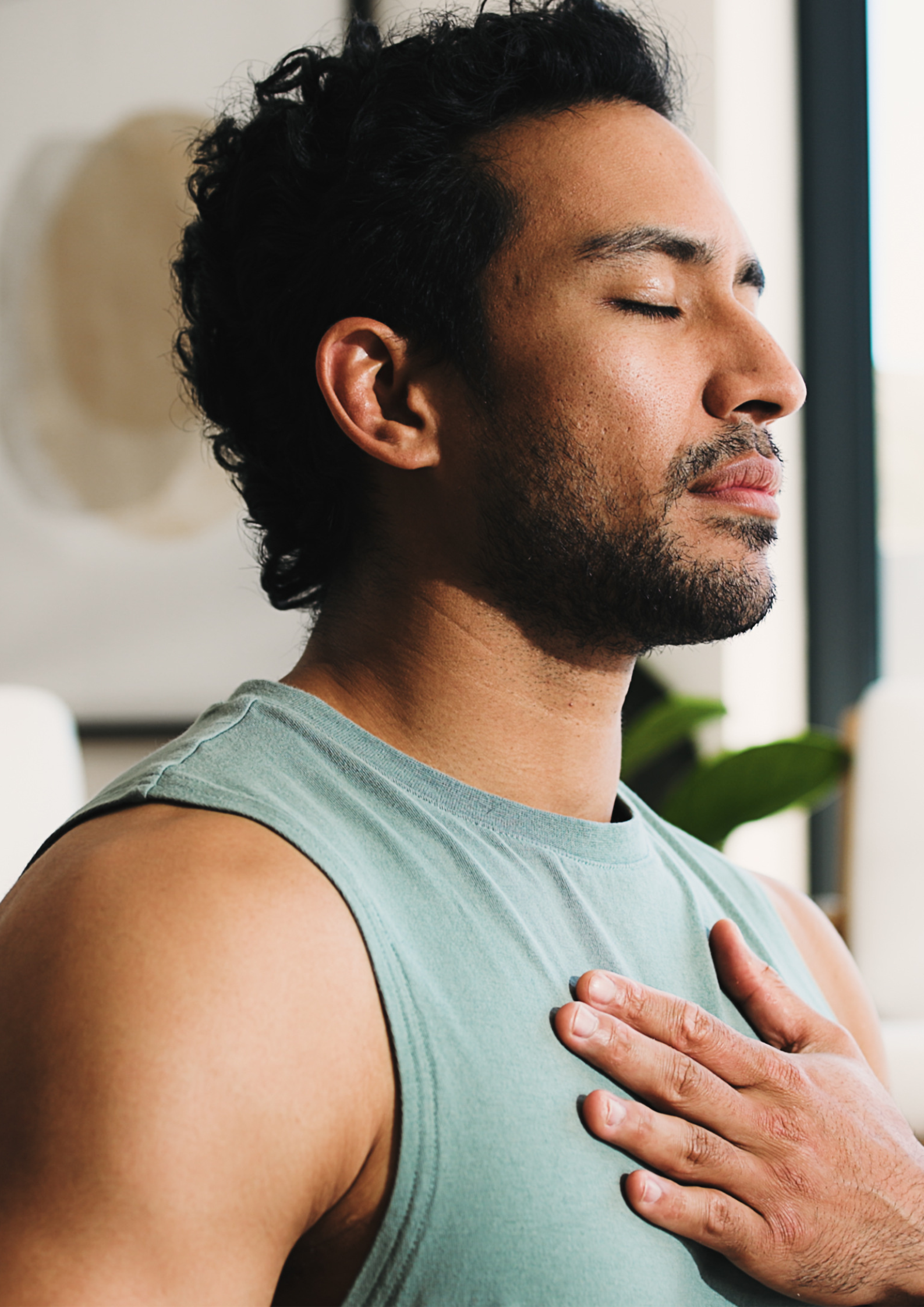 A man with closed eyes and a beard places his hand over his heart during a moment of reflection or meditation indoors with natural light and a plant in the background.