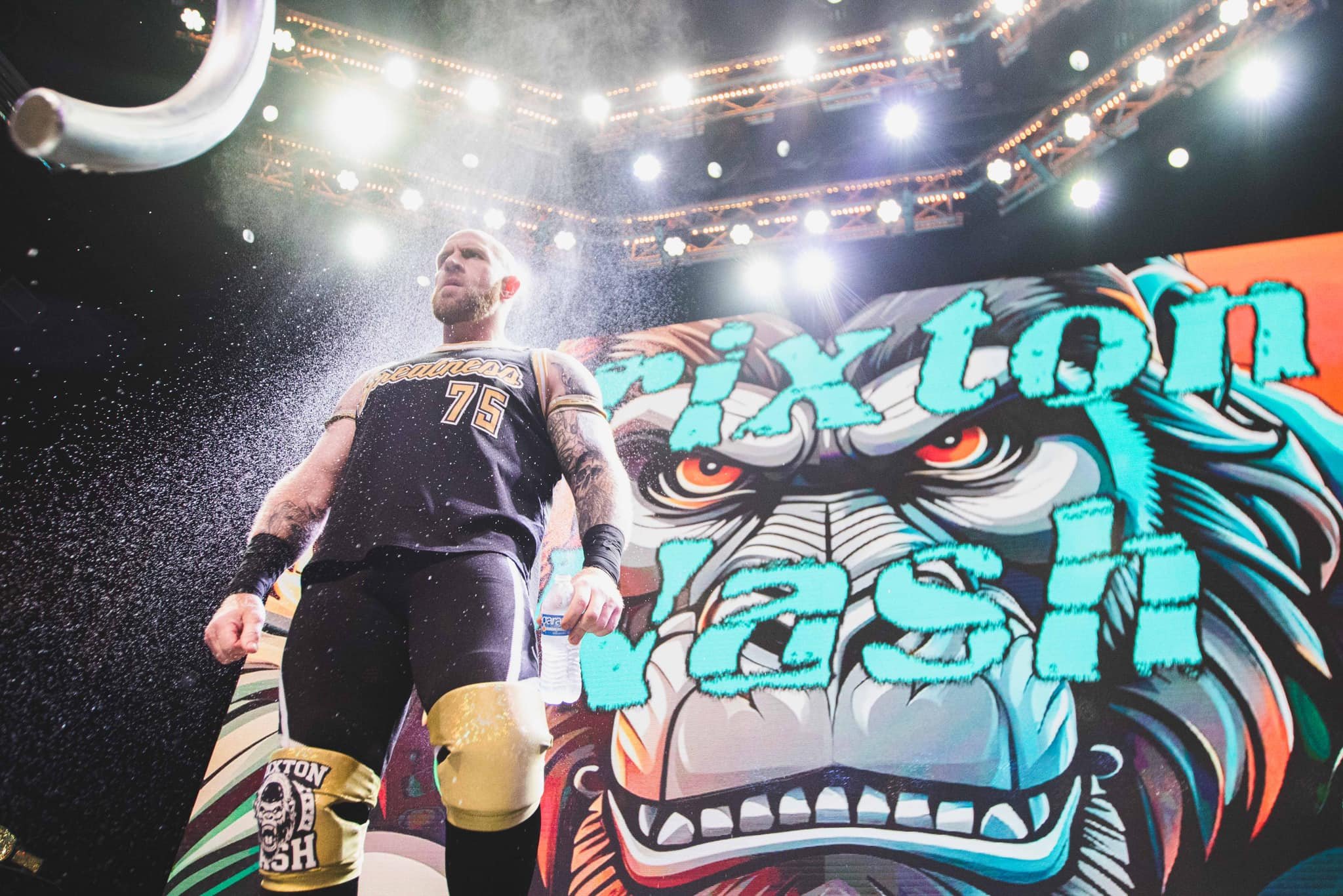 A wrestler in black and yellow gear standing in front of a colorful tiger mascot graphic with the words 'Pix Con' on it at a wrestling event, water being sprayed over him.