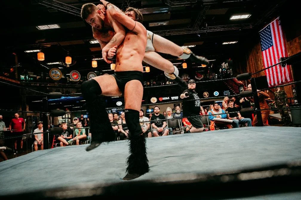 Two wrestlers engaged in a match inside a wrestling ring with an audience watching. One wrestler is lifting the other.