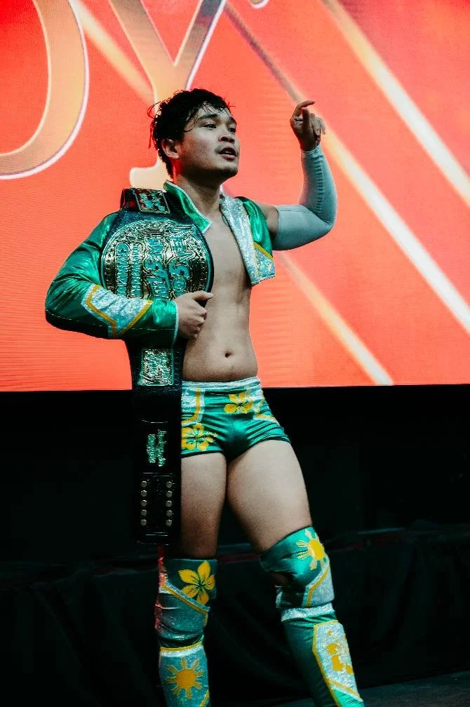 A male professional wrestler standing on stage, wearing vibrant green and yellow wrestling gear with floral patterns, holding a championship belt over his shoulder, and posing with one arm raised.