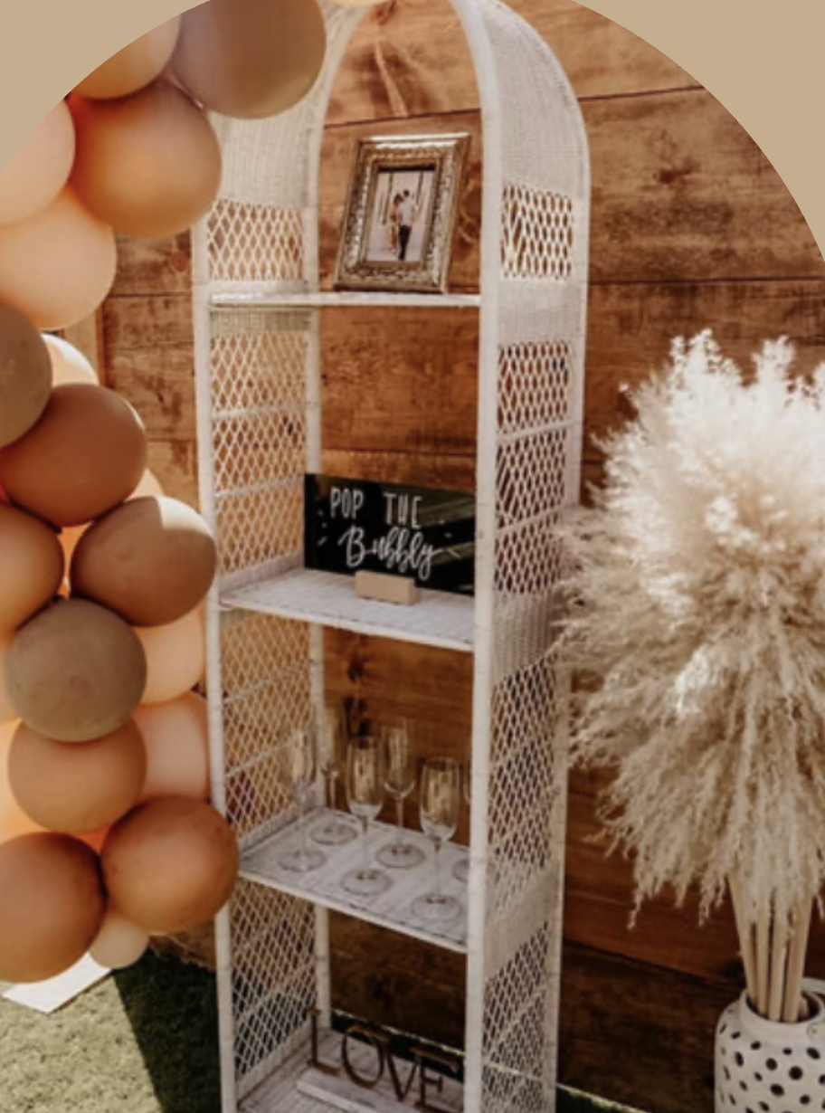 Decorative white shelving unit with four shelves, placed against a wooden wall, holding a framed photo, a sign reading 'Pop the bubbly,' glasses, and a 'LOVE' sign, with a large dish of pampas grass in a white vase next to it.