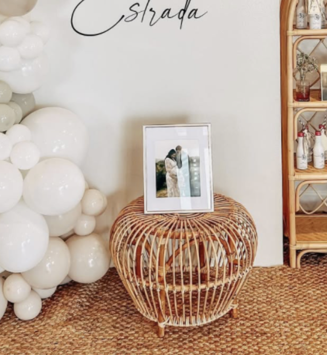 A wicker side table with a framed photograph of a couple on top, next to a ball sculpture made of white spheres, and a wooden shelving unit with decorative bottles on the right.