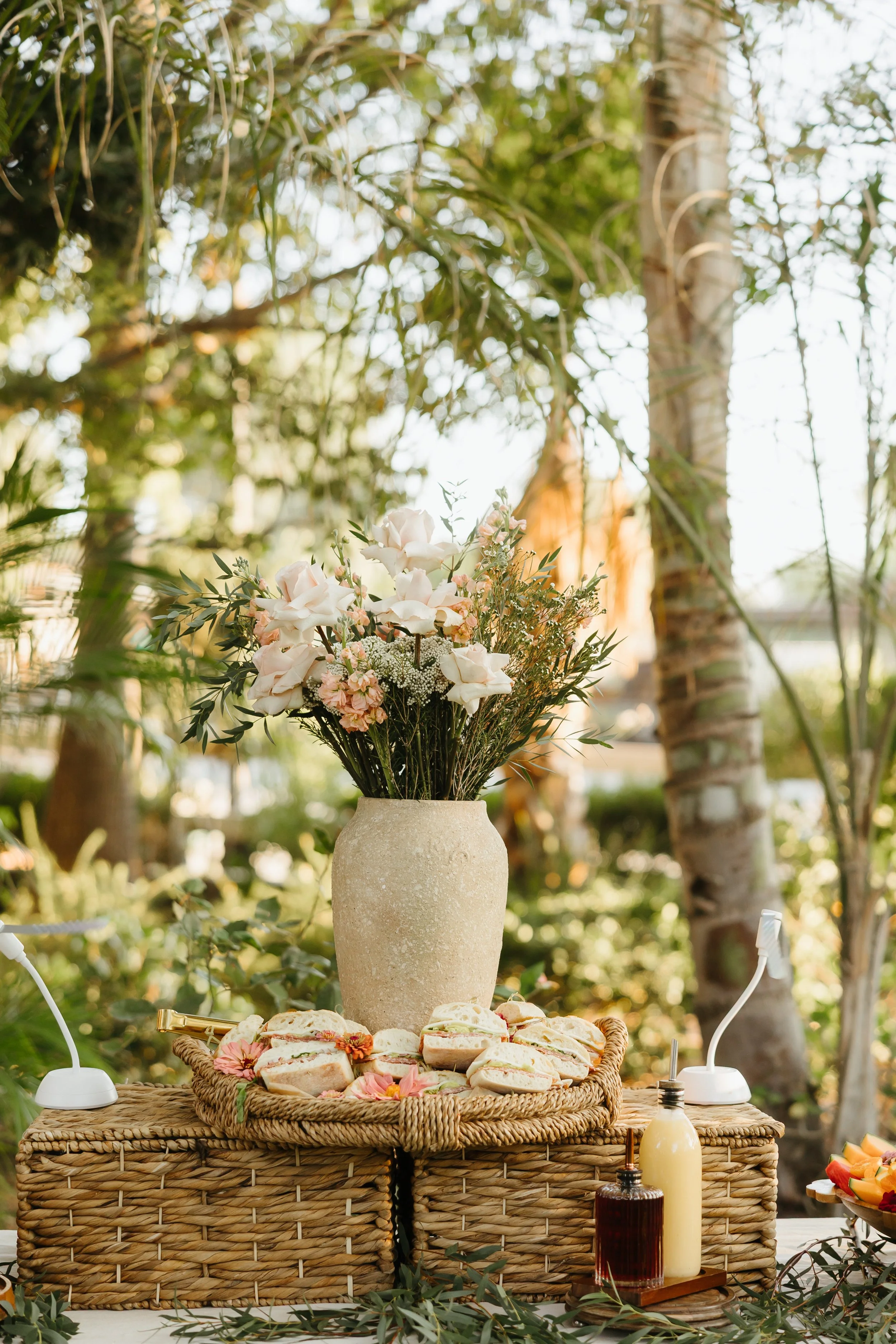 A floral centerpiece with roses and greenery in a beige vase, placed on a wicker basket with sandwiches, surrounded by bottles and electric cords, set outdoors with trees and sunlight.
