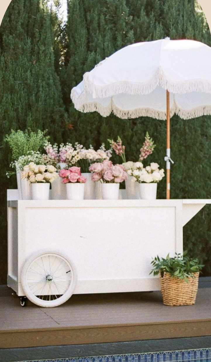 A white flower cart with a large white parasol, filled with white and pink flowers in white pots, and a wicker basket with greenery on the side, set outdoors against a backdrop of tall green trees.