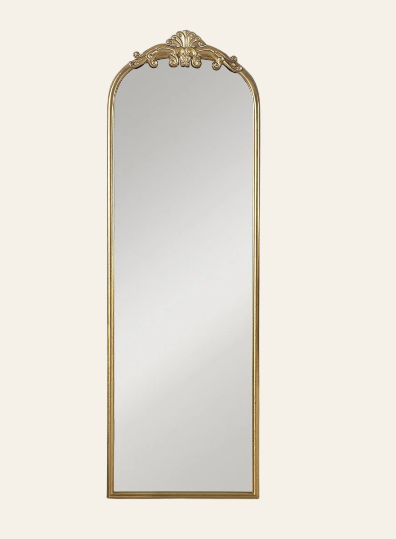 Decorative full-length mirror with a gold ornate frame and arched top against a light background.
