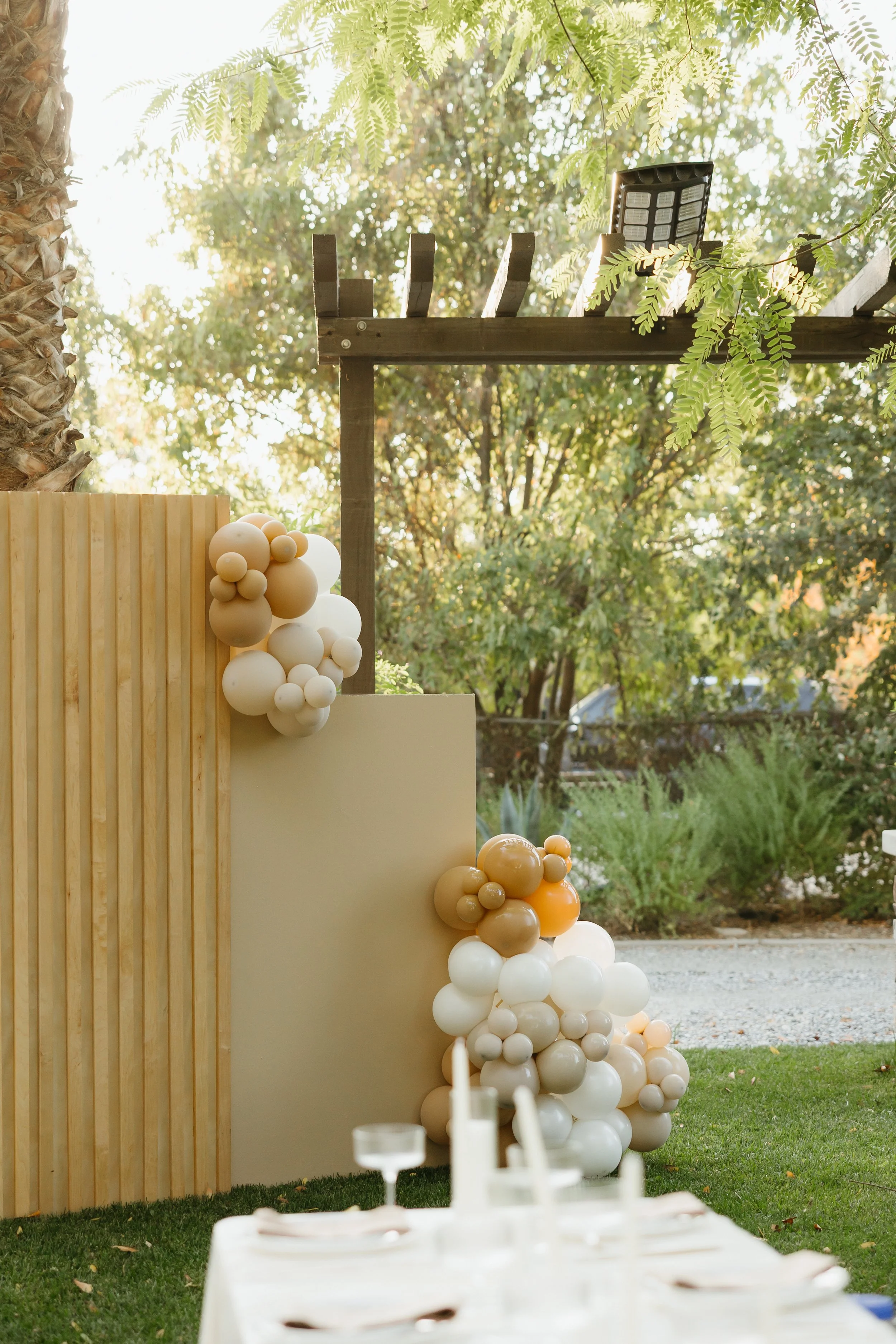 Decorative balloon arrangement in beige, white, and orange tones outside near a wooden wall with greenery in the background and a table set for an outdoor event.