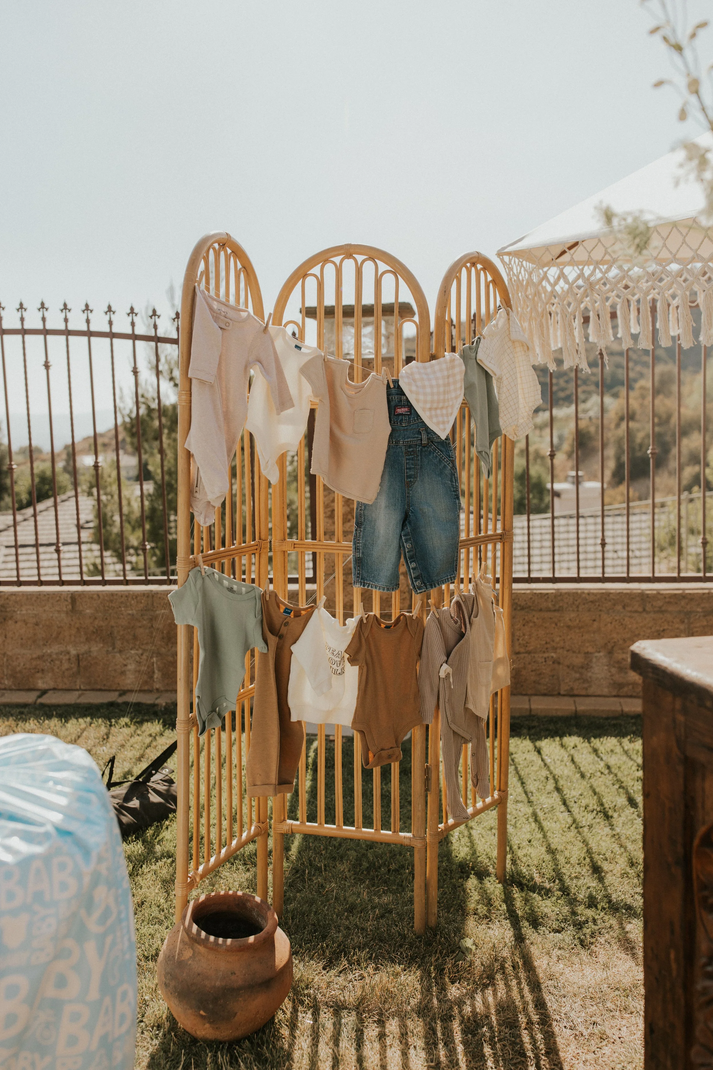 Baby clothes hanging on a bamboo room divider on a patio, with a white umbrella and a view of rooftops and hills in the background.