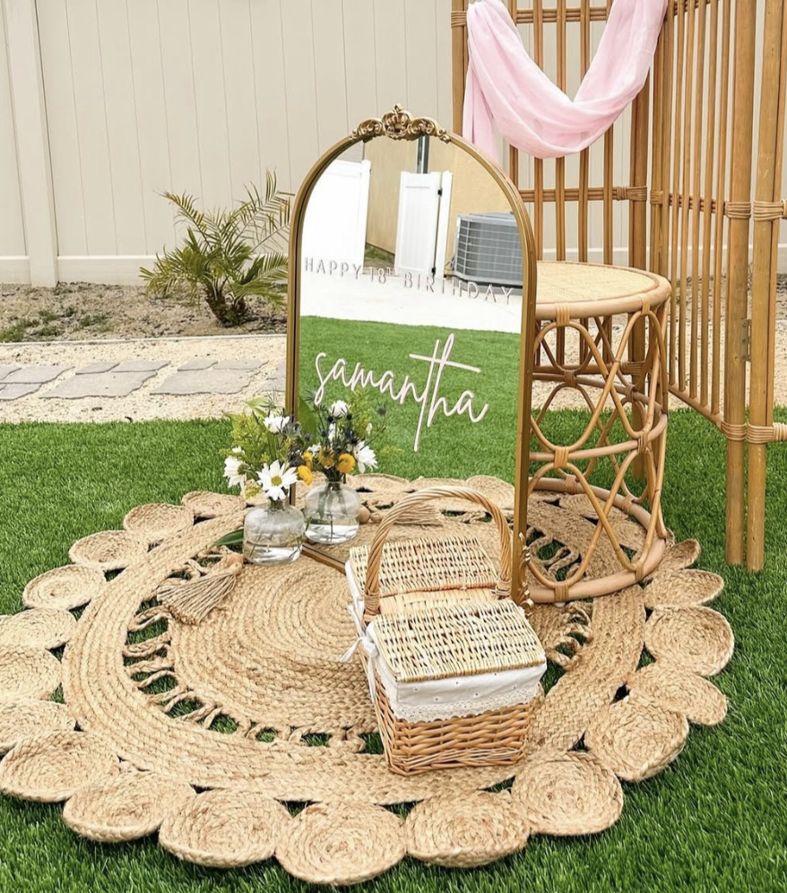 Decorative outdoor setting with a large mirror that has the name 'Samantha' written on it, two small vases of flowers, two wicker baskets, and a rattan umbrella stand on a circular woven rug on grass. In the background, there is a bamboo structure with a pink cloth draped over it, a small tree, and a beige wall.