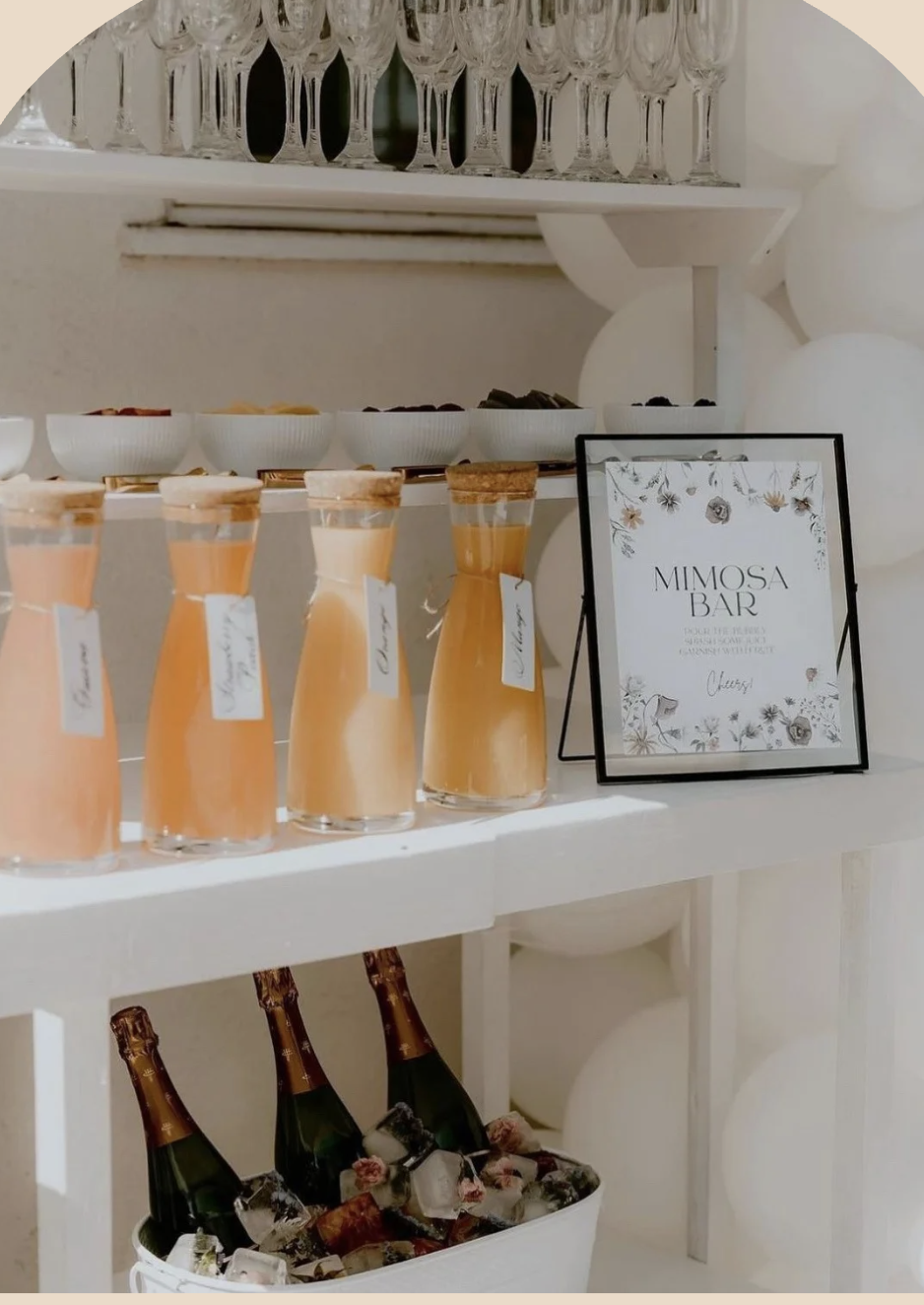 A mimosa bar with bottles of orange juice, bowls of fruit, and a sign that reads 'MIMOSA BAR' with floral decorations, set up for a celebration or event.