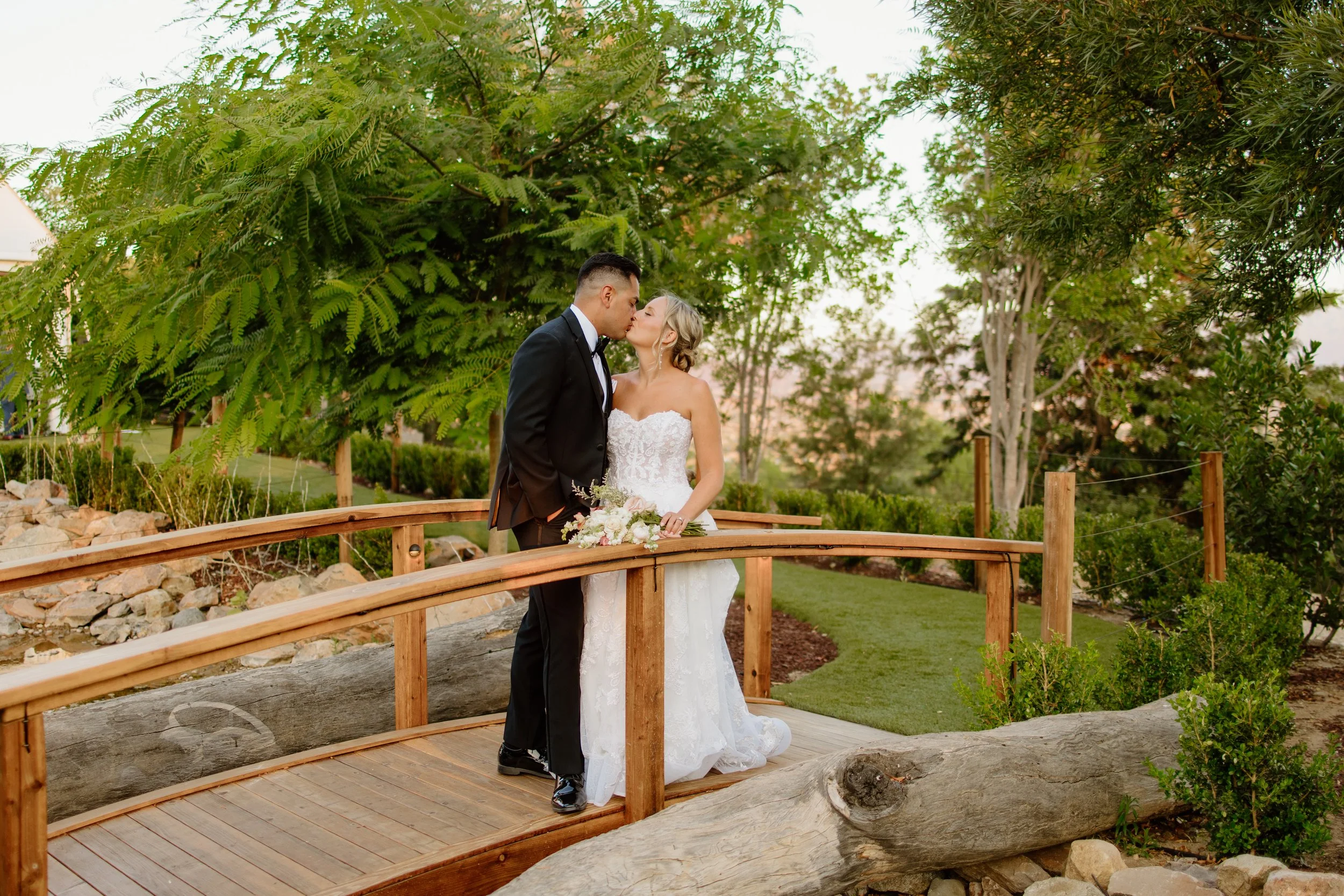 A bride and groom share a kiss on a wooden bridge in a park, surrounded by green trees and plants at sunset.