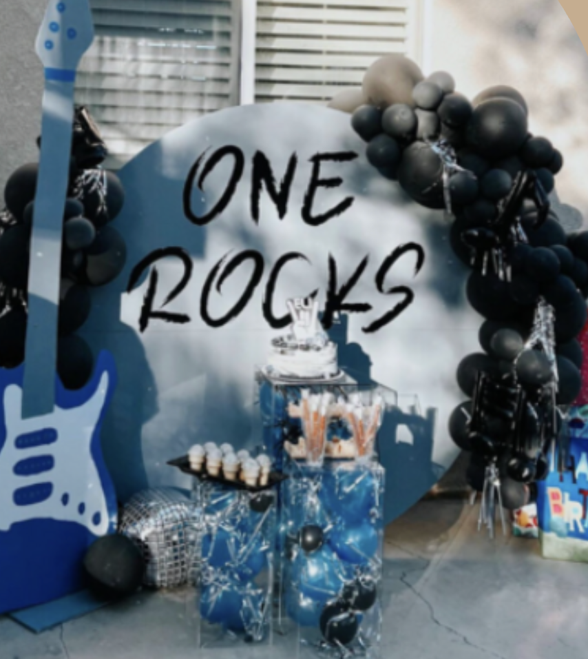 Celebration setup with a large sign that reads 'ONE ROCKS,' black and gray balloons, a table with cake pops and treats, and a blue and white guitar decoration.
