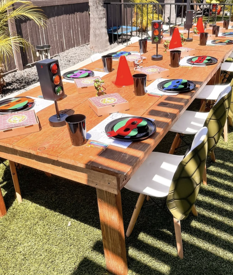 Outdoor birthday party table set with black, red, green, and purple masks, traffic light centerpieces, pizza boxes, black cups, and party hats, with sunlight and chairs around the table.
