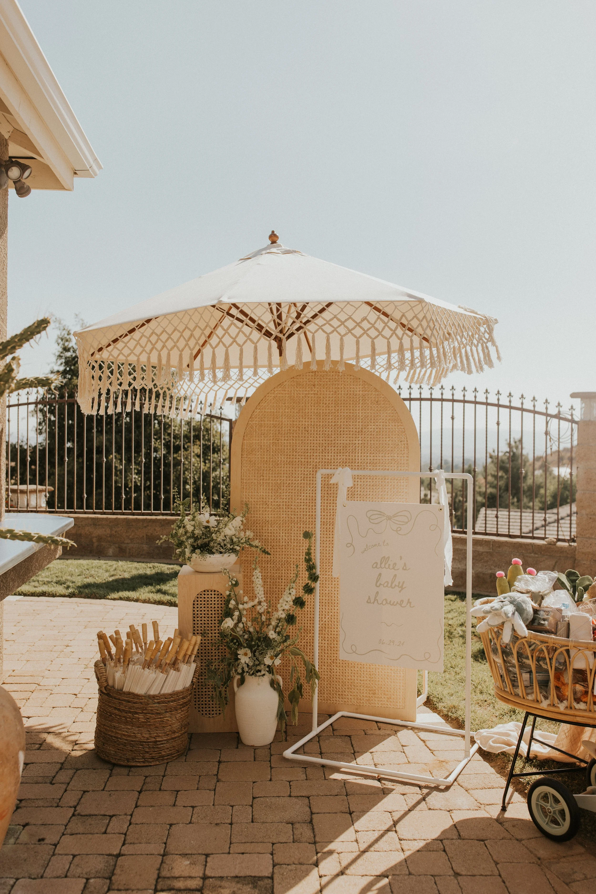 Outdoor baby shower setup with a cream-colored umbrella, a whiteboard sign, flower arrangements, and gifts, on a patio with a brick ground and a metal fence in the background.