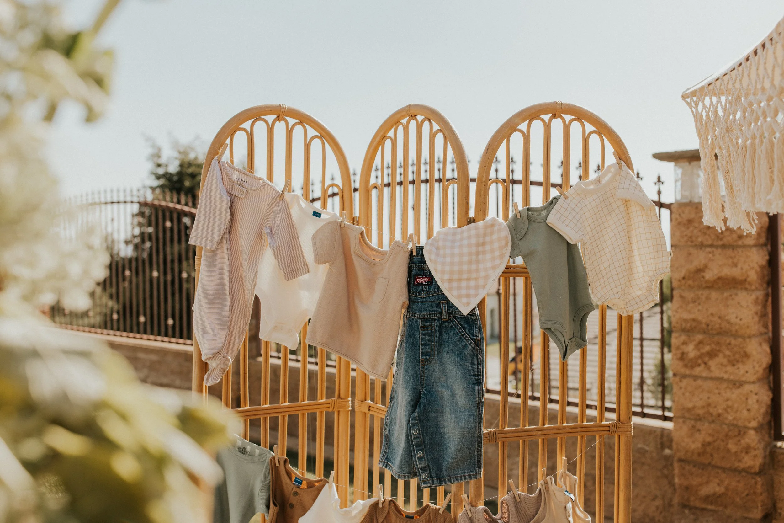 Baby clothes hanging on a wooden room divider outdoors, including onesies, a skirt, a heart-shaped cloth, and a checkered shirt.