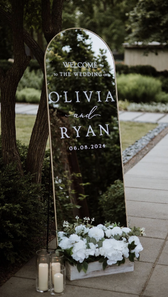 A glass wedding sign with white lettering reading 'Welcome to the wedding of Olivia and Ryan, 06.08.2024,' placed next to white floral arrangements and candles on a sidewalk in an outdoor garden setting.
