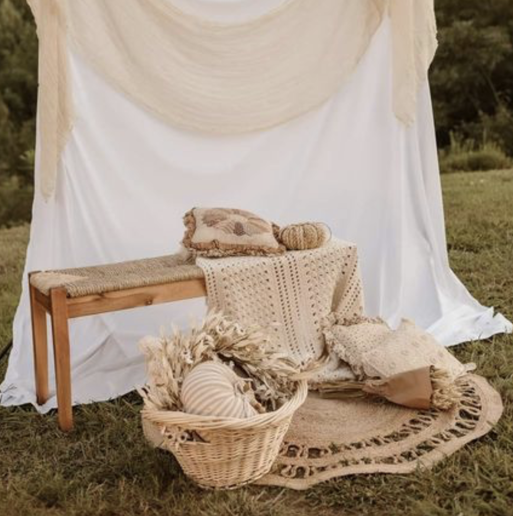Bohemian-themed outdoor scene with a bench, cushions, rugs, and a basket of dried decorative items, arranged in front of a white draped fabric backdrop in a grassy area.