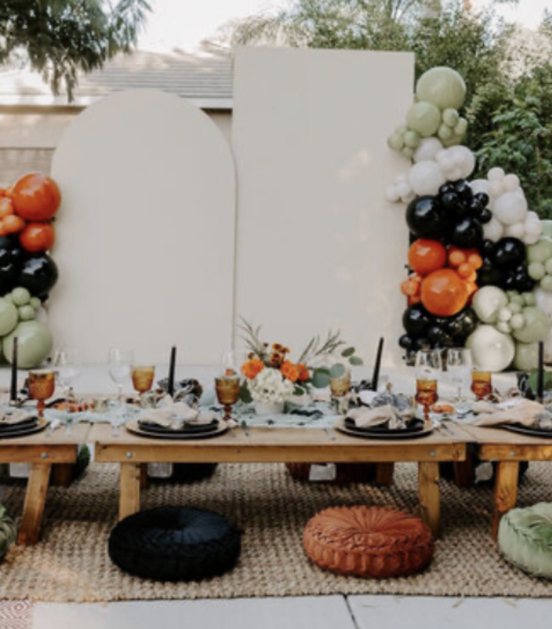 Outdoor dining table with fall-themed decorations, surrounded by colorful pillows, and a balloon arch featuring black, white, orange, and green balloons, with a white backdrop and greenery in the background.