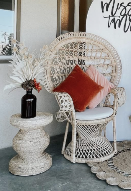Decorative wicker chair with two pillows, one red and one pink, next to a textured side table with a dark vase holding dried white and beige plants. Part of a sign with black text is visible in the background.