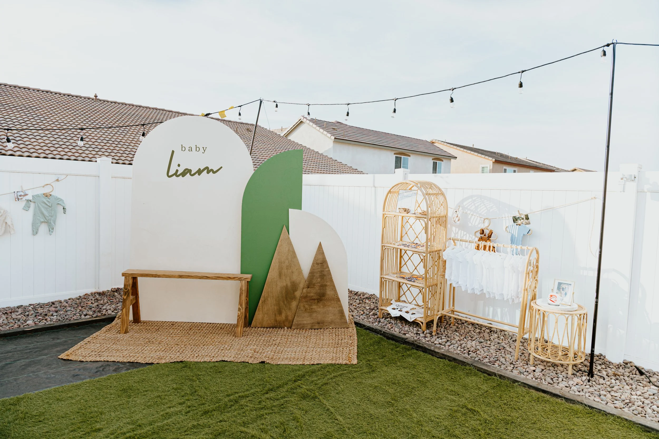 Decorative display for a baby shower with a backdrop reading 'baby Liam,' wooden and wicker furniture, and baby clothes hanging on a line in a backyard with a white fence and string lights.
