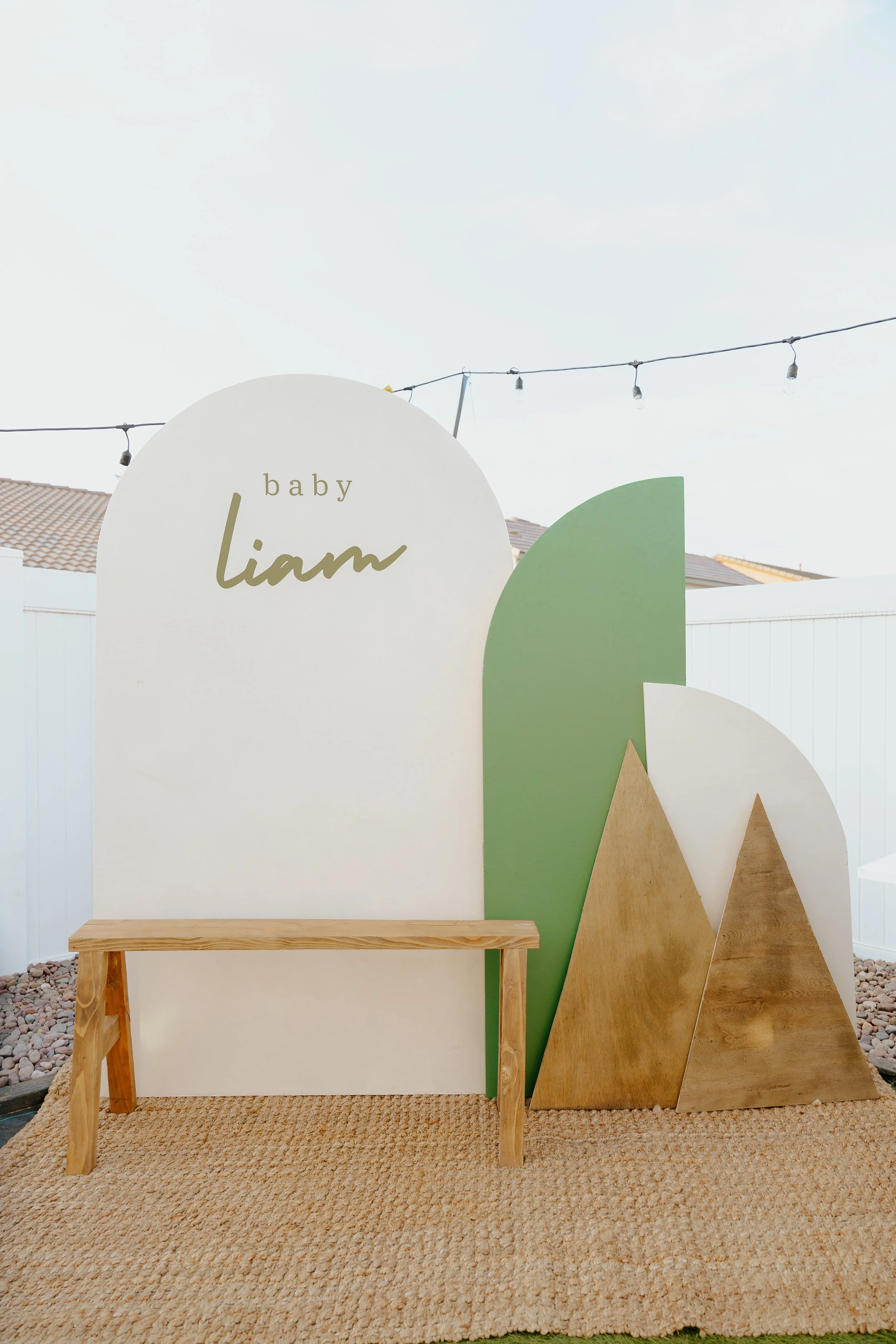 Decorative backdrop for a first birthday celebration with mountains and leaf shapes, bearing the words 'baby Liam' written on the white mountain, and a small wooden bench in front.