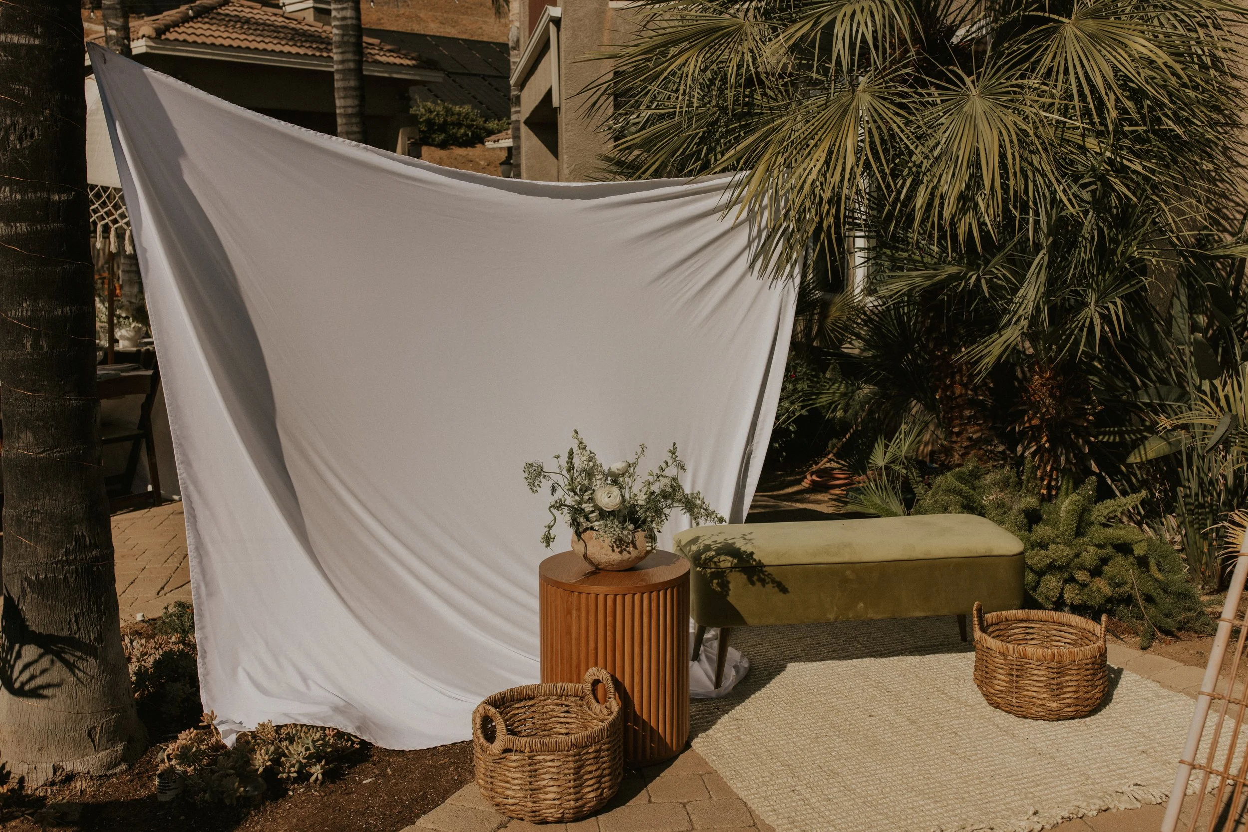 Outdoor scene with a white fabric backdrop, a small wooden table with a floral arrangement, a green cushioned bench, wicker baskets, a beige rug, and lush green plants and palm trees.
