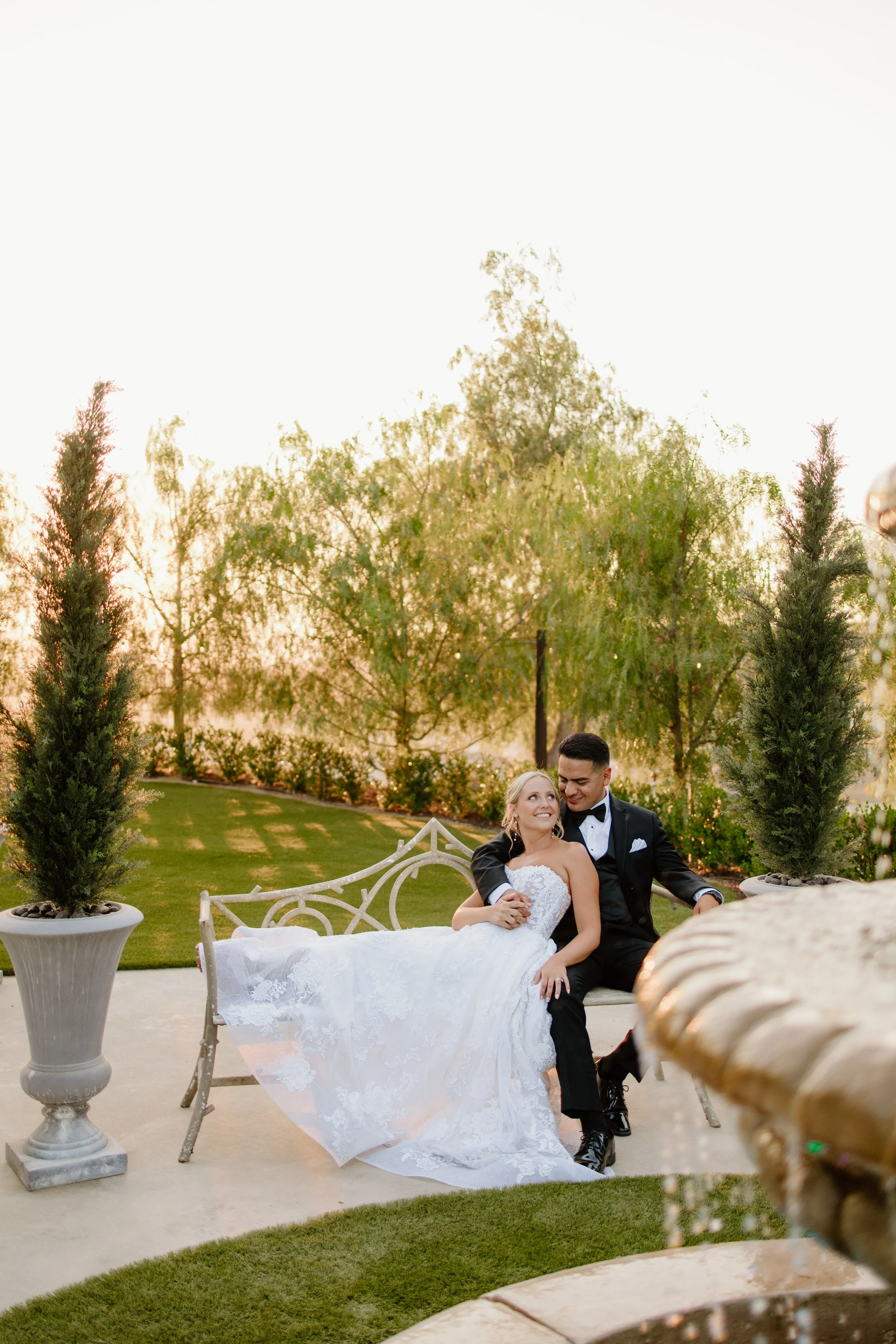A newlywed couple sitting on a white ornate bench outdoors during sunset, surrounded by potted trees and greenery, with a stone table partially visible in the foreground.
