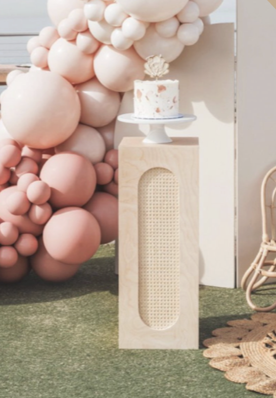 A small white cake with a flower on top, placed on a cake stand, on a wooden pedestal, with pink and beige balloon decorations behind it.