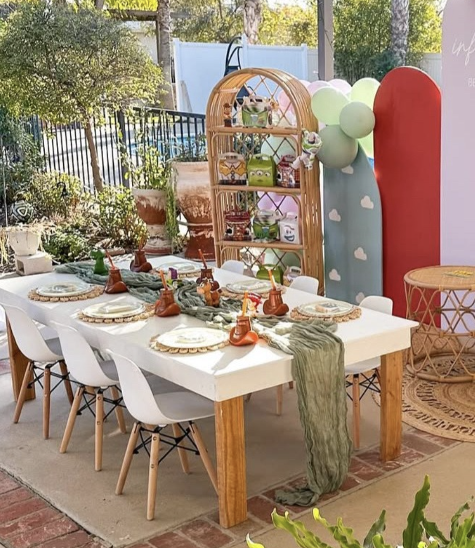 Decorated outdoor area for children's party with a long white table set with plates and drink dispensers, surrounded by white chairs. Behind the table, there's a colorful backdrop with clouds, balloons, and a wicker shelf with toys and books, and plants on the porch.