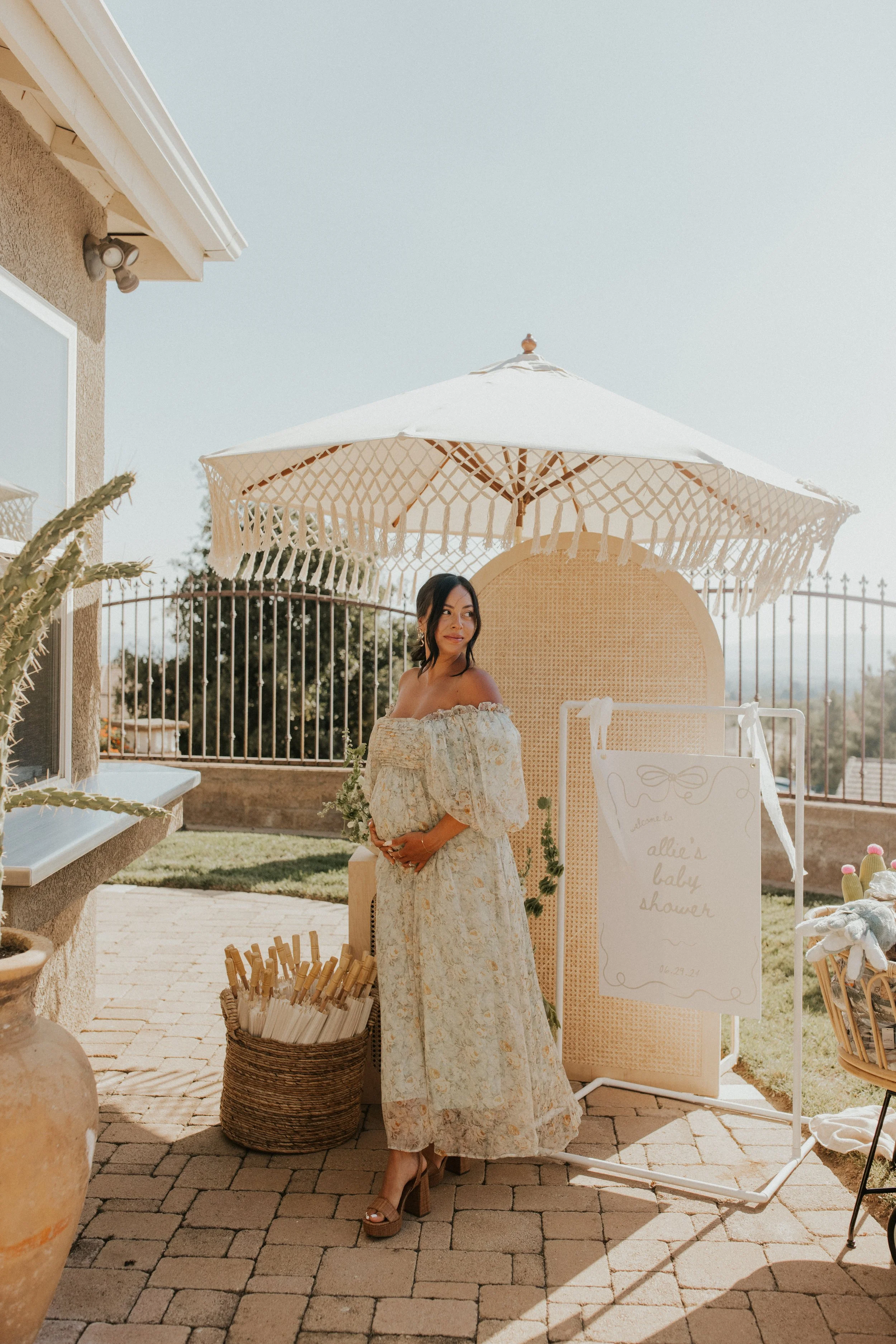 A pregnant woman standing outdoors under a large patio umbrella, wearing a long floral dress and heels, with a welcome sign for a baby shower that reads 'Welcome to allie's baby shower' in elegant script.