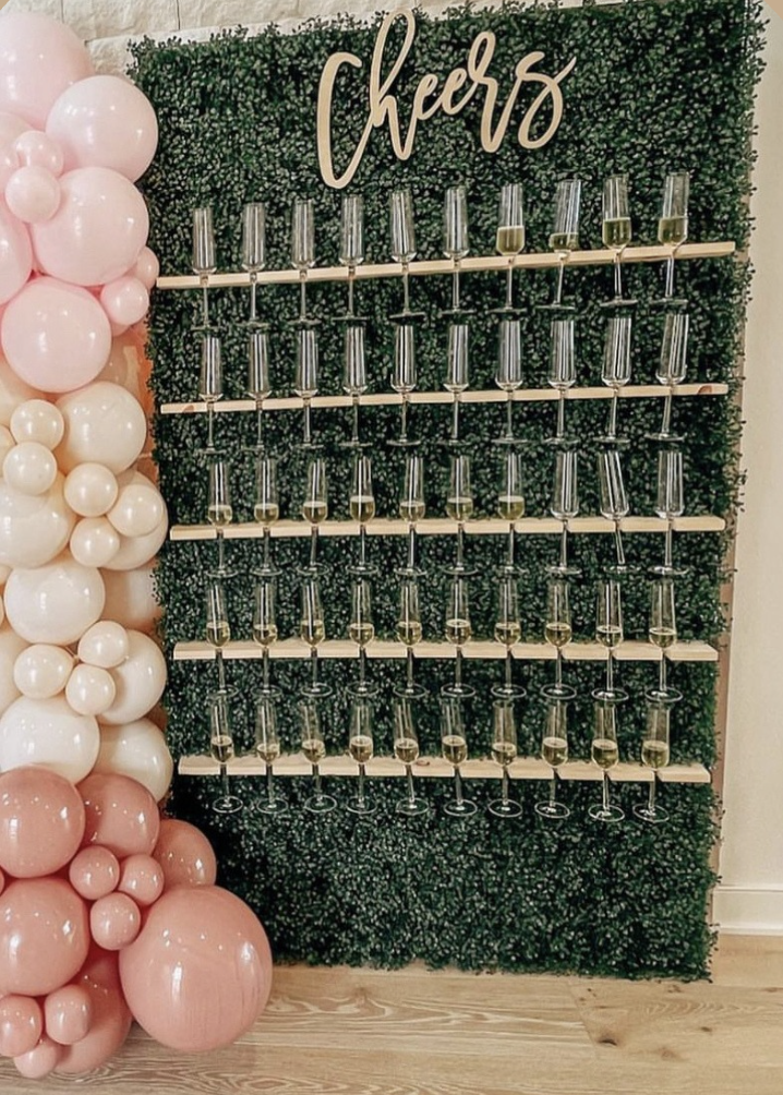 Decorative photo booth with a green foliage backdrop, a 'Cheers' sign at the top, and rows of empty champagne glasses on shelves. To the left, there are pink and white balloons arranged in a cluster.
