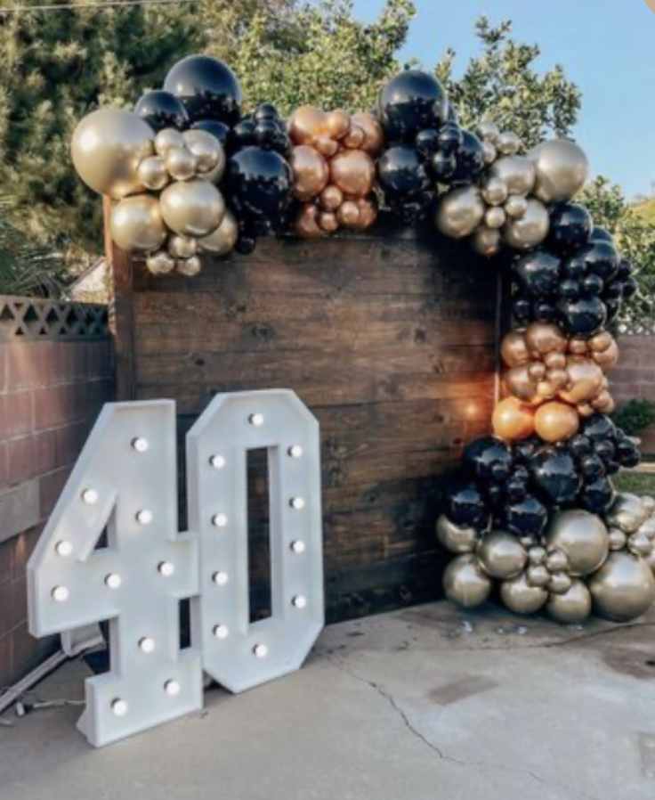 Decorative birthday display with a large illuminated number 40, a wooden backdrop, and a balloon arch with black, gold, and copper balloons.