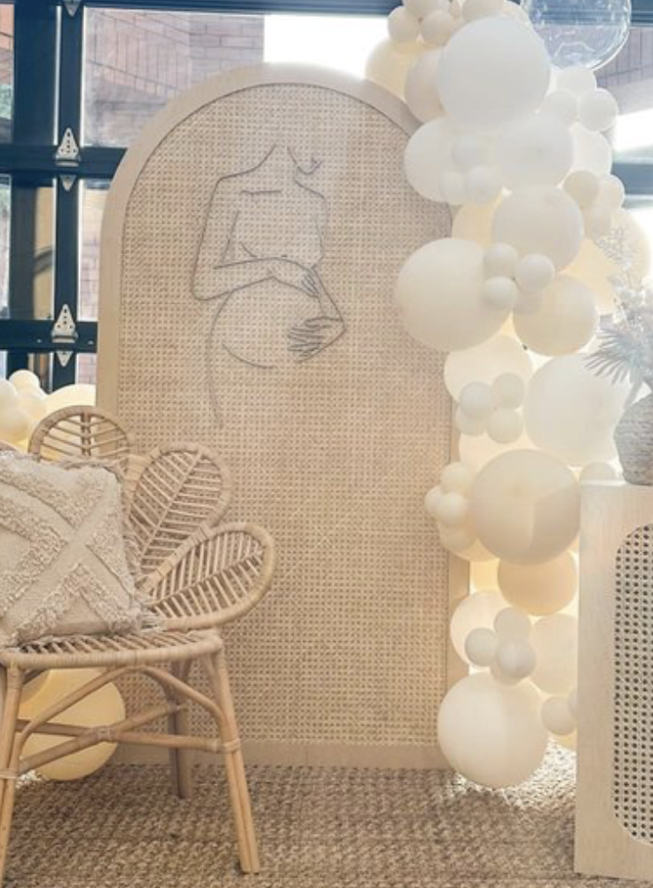 Decorative indoor space with a rattan chair, a cushion, a beige woven room divider with a line drawing of a pregnant woman, and white balloons.