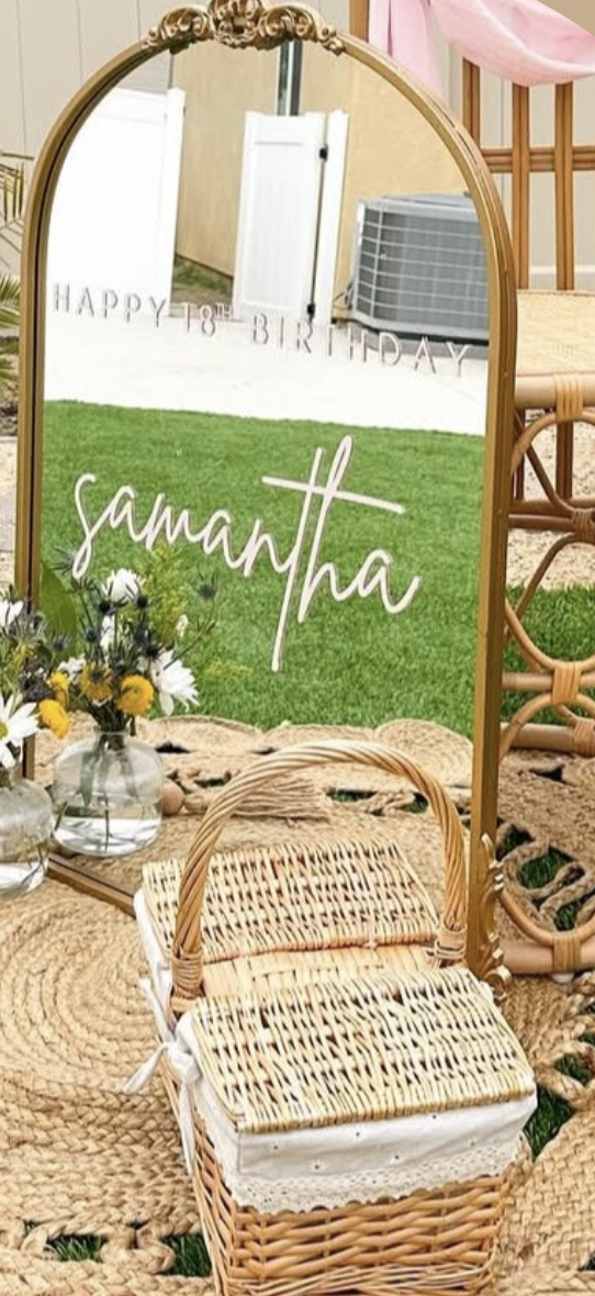 Mirror with 'Happy 18th Birthday Samantha' written on it, surrounded by flowers, wicker chairs, and a woven rug, indicating a birthday celebration setup.