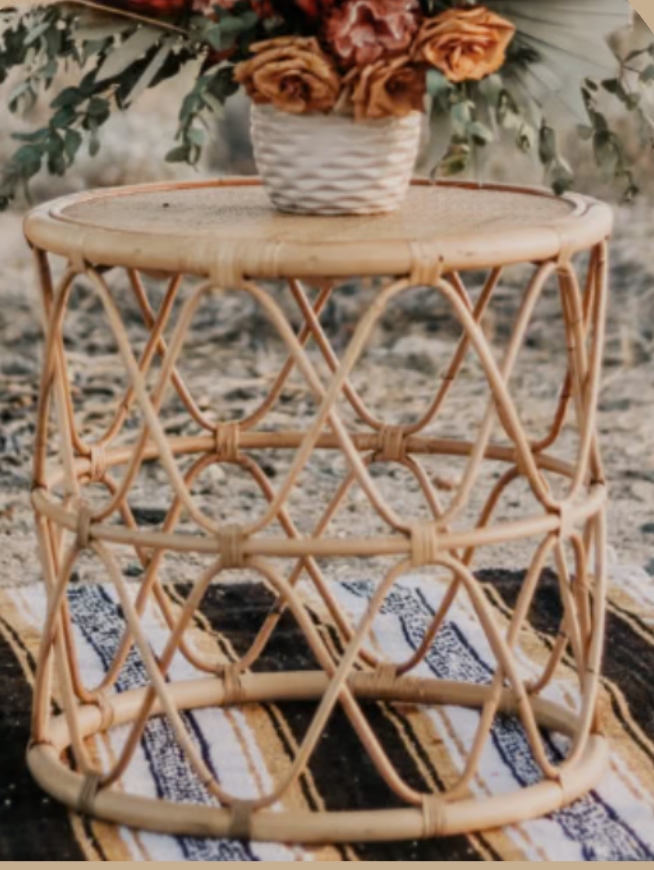 A round wicker side table with an intricate lattice design, placed on a patterned rug, with a white woven basket of flowers on top. The background is a neutral outdoor setting.