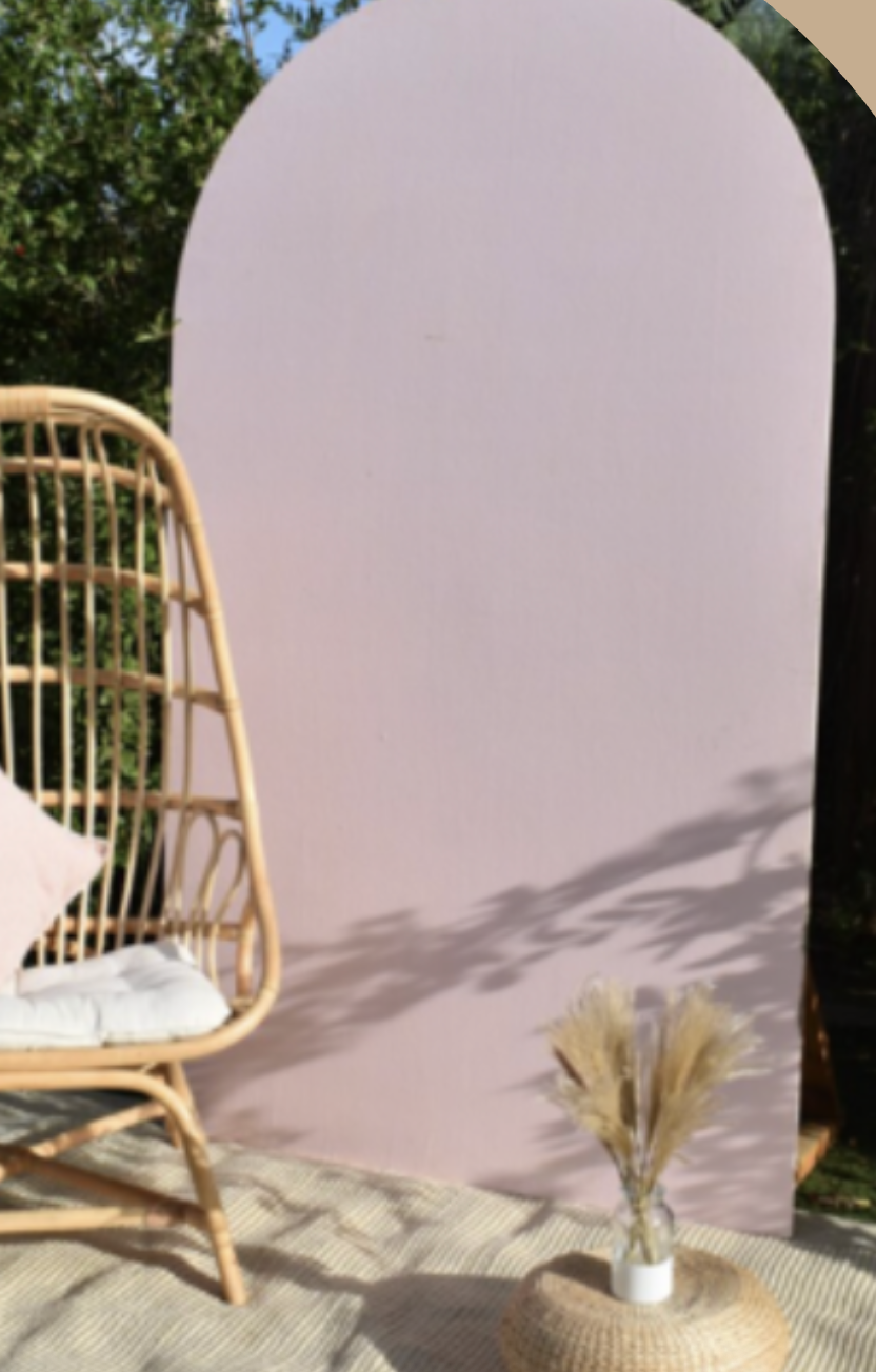 An outdoor scene with a rattan chair, a pink panel backdrop, and a vase with dried pampas grass on a woven mat, set against trees and greenery.