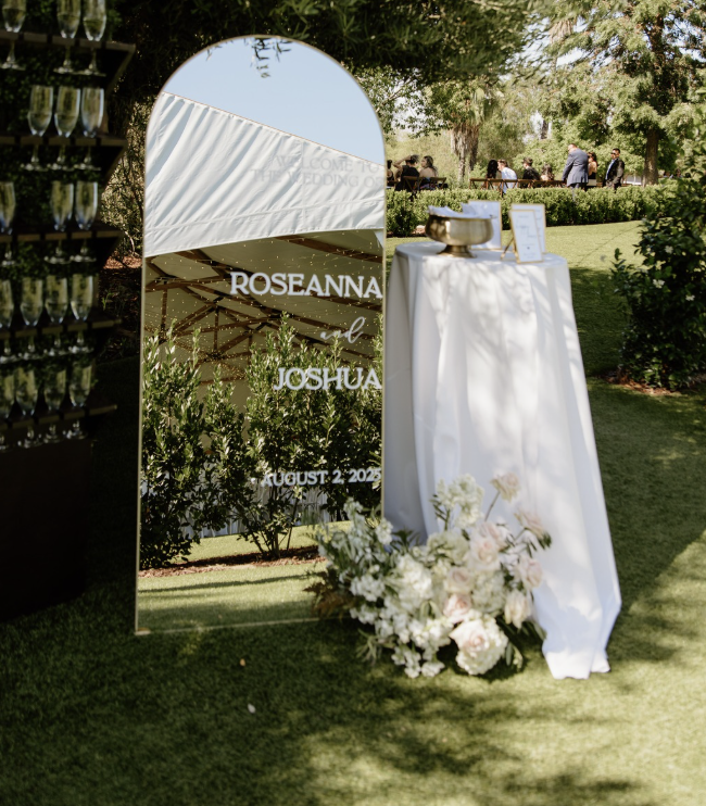 A wedding reception area outdoors with a mirror displaying the names Roseanna and Joshua, and the date August 2, 2023. There's a table with a white cloth, a flower arrangement, and a bowl, with guests in the background.