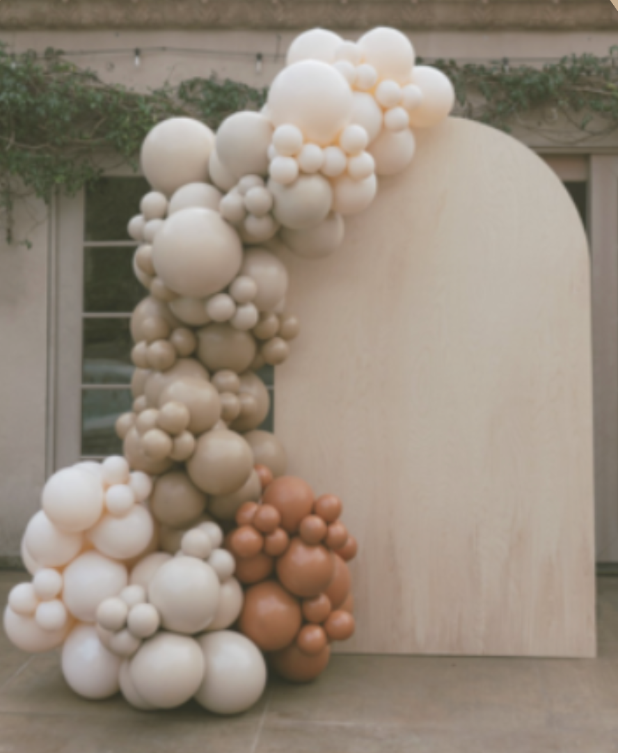 Decorative balloon arrangement with white, beige, and brown balloons next to a cream-colored, curved wall backdrop.