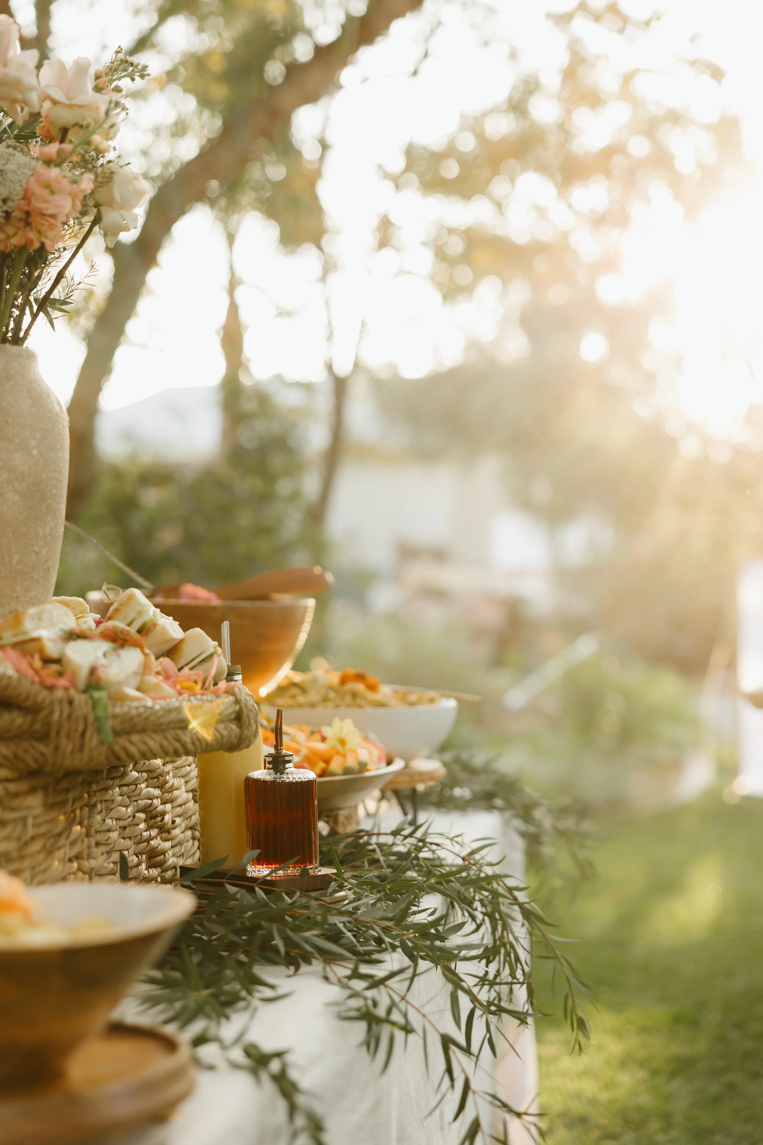 Outdoor buffet table with bowls of food and condiments, decorated with green foliage, set in a natural outdoor setting with trees and sunlight in the background.