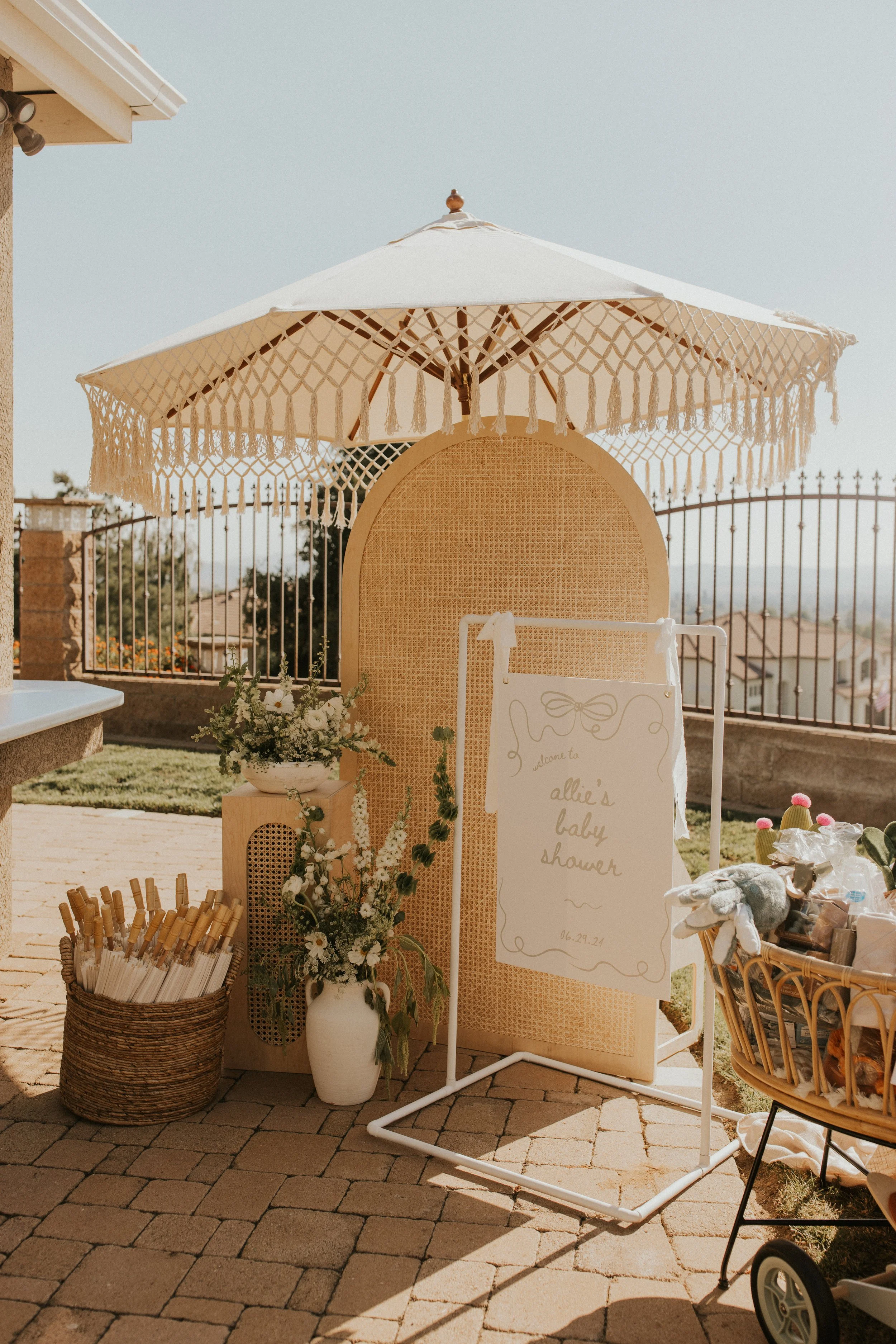 Decorative setup for a baby shower with a large parasol, flowers, a sign that reads 'Allie's baby shower', and various gifts and supplies.