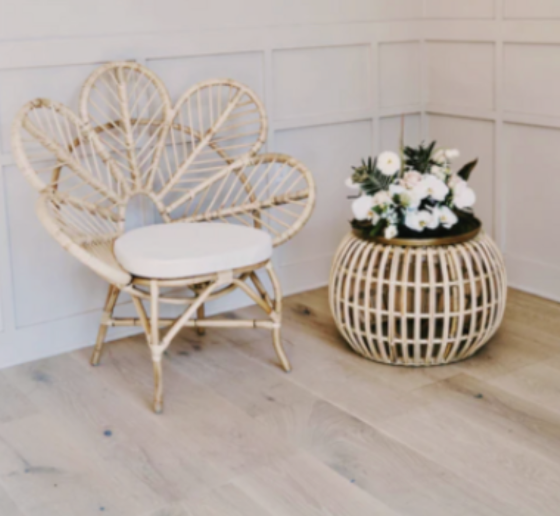 A wicker chair with a white cushion next to a round wicker table holding a bouquet of white flowers, against a white paneled wall with wooden flooring.