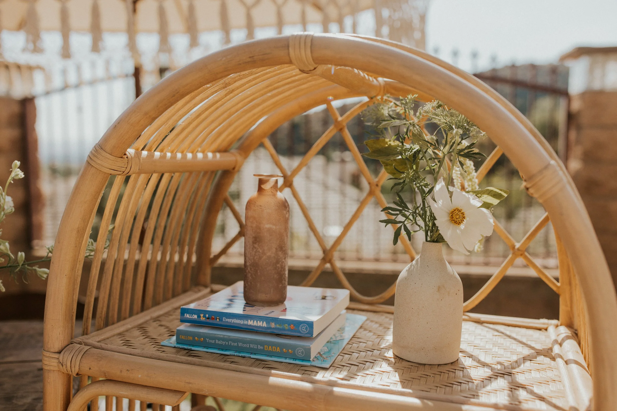 A wicker outdoor chair with a woven seat, holding a stack of books, a small weathered brown bottle, and a white ceramic vase with white flowers and greenery, on a balcony or patio with wooden fencing.