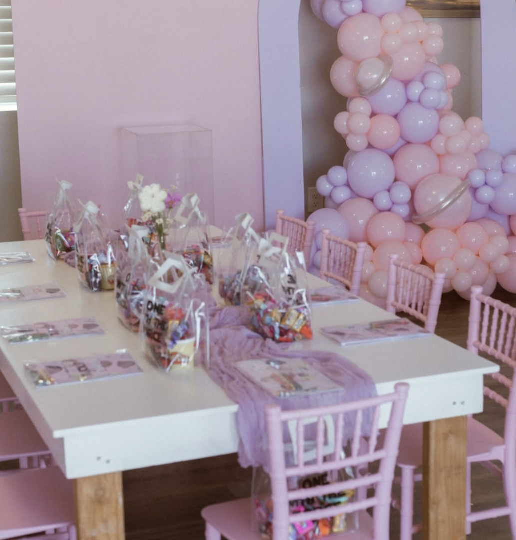 Party table decorated with pink balloons in the background, pink chairs, and gift bags on the table, indicating a celebration for a young girl.