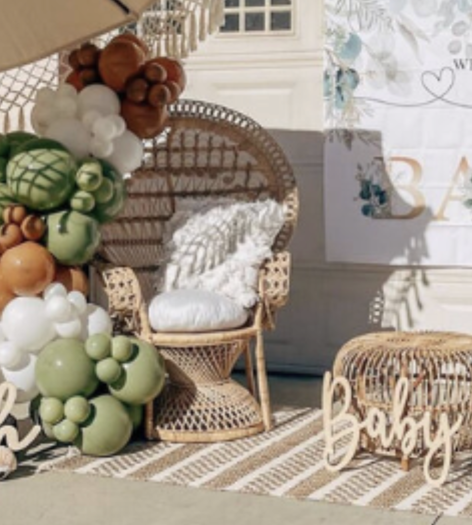 Decorative area with wicker furniture, including a chair and a small ottoman, surrounded by pastel-colored balloon arrangements, a striped rug, and a white sign with floral accents and the word 'BABY'.