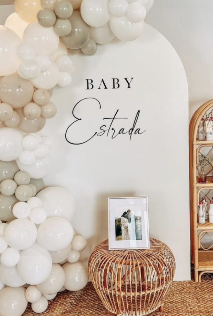 Decorative setup with a white wall, balloon arch with white, beige, and gray balloons, a framed photo of a woman and a child, a wicker table, and a bamboo shelf with bottles and decorations, with the text "Baby Estrada" on the wall.