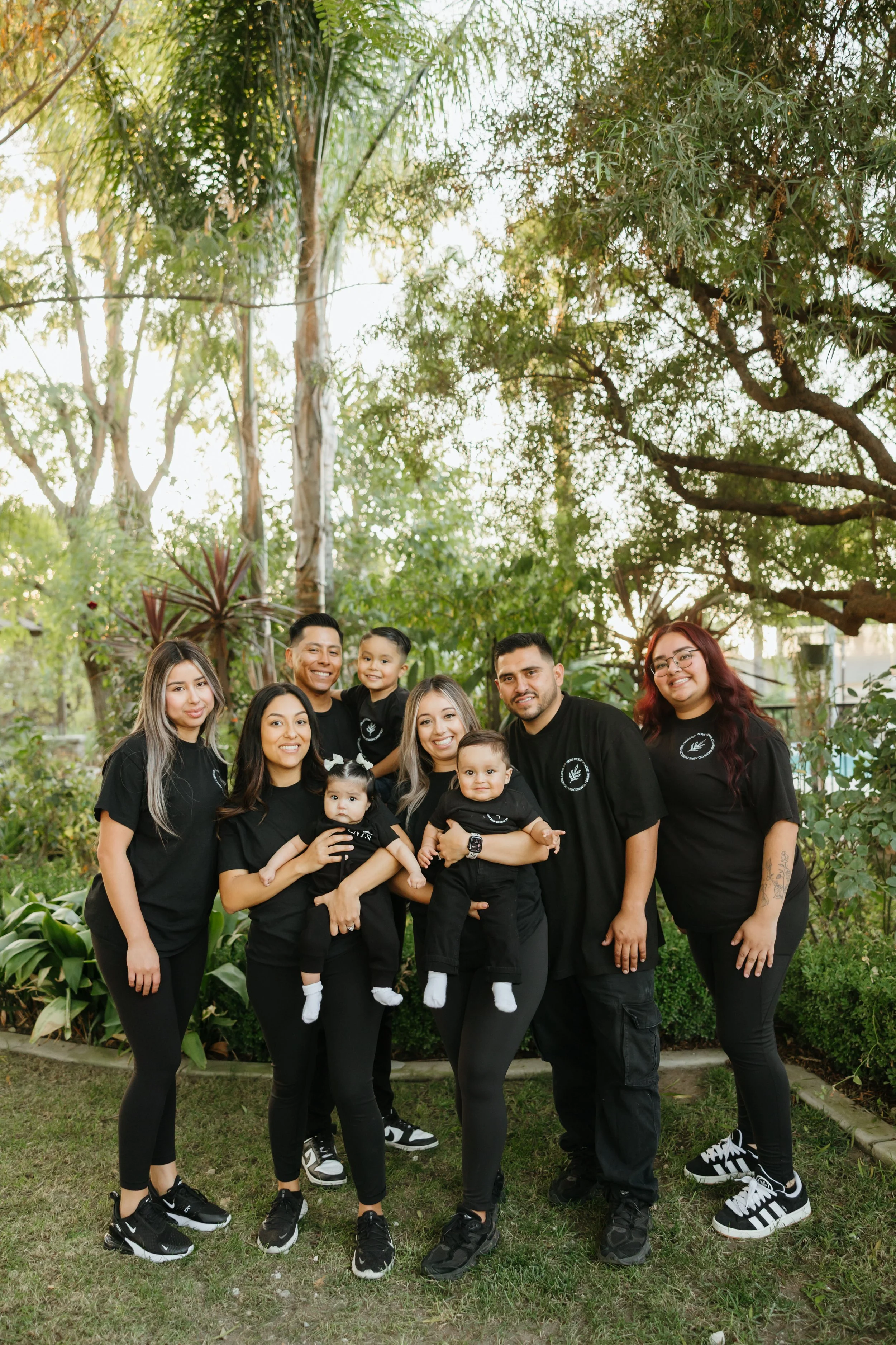 Family gathering outdoors with nine members, including adults and children, all wearing matching black outfits, standing on grass with trees and plants in the background.