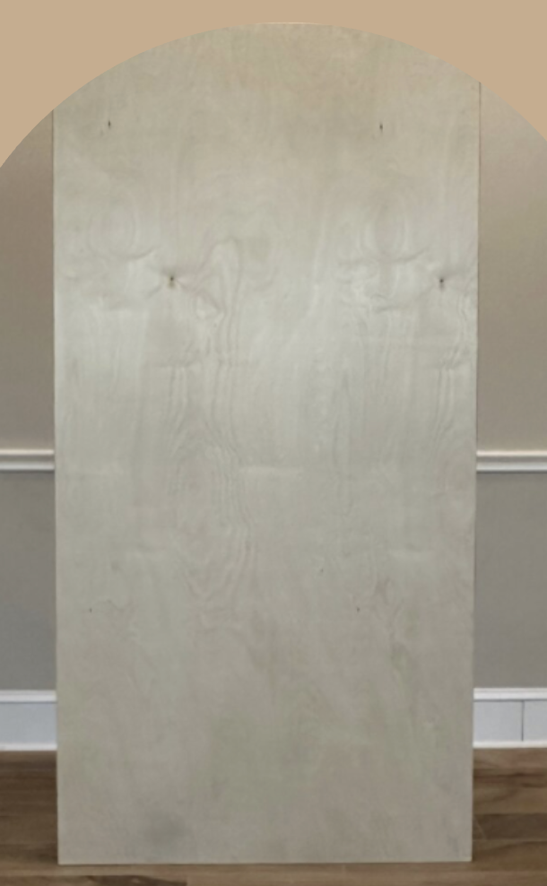 A plain piece of light-colored wood with two small buttons or knots and a rounded top, leaning against a beige wall with white trim and a wooden floor.