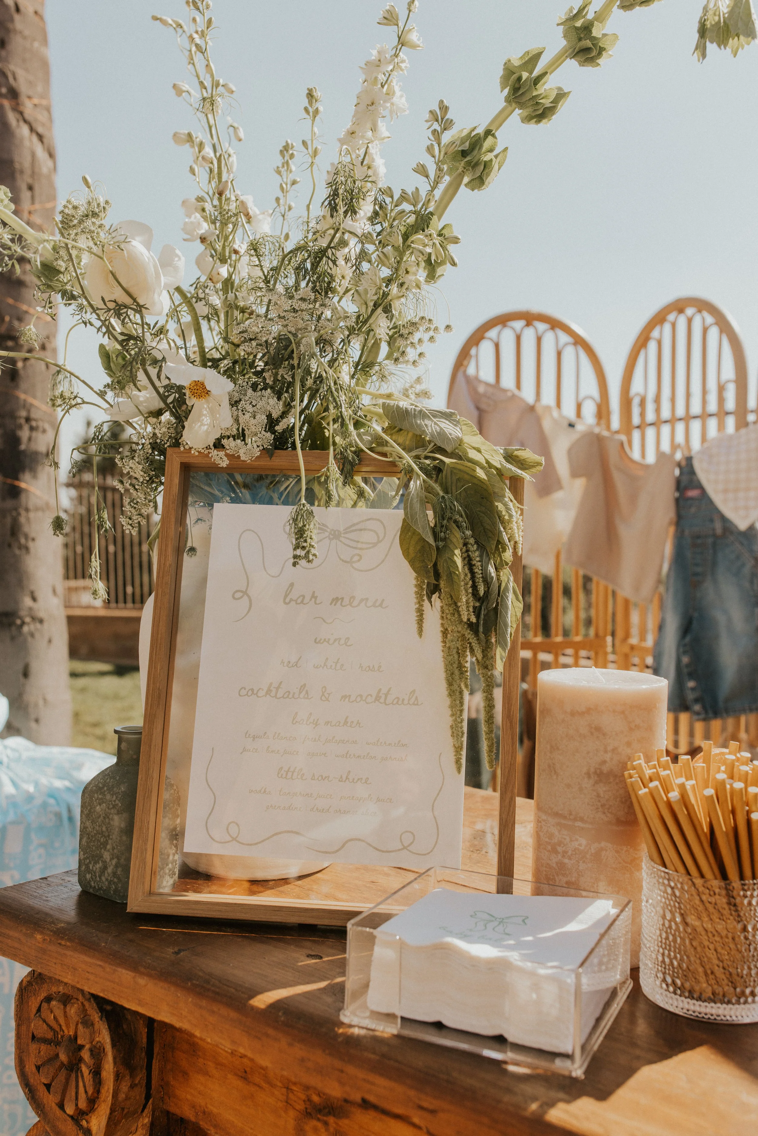 A rustic outdoor bar station decorated with a floral arrangement, a bar menu sign, a candle, a container of straws, and a small stack of napkins. In the background, clothes are hanging on a wooden fence.