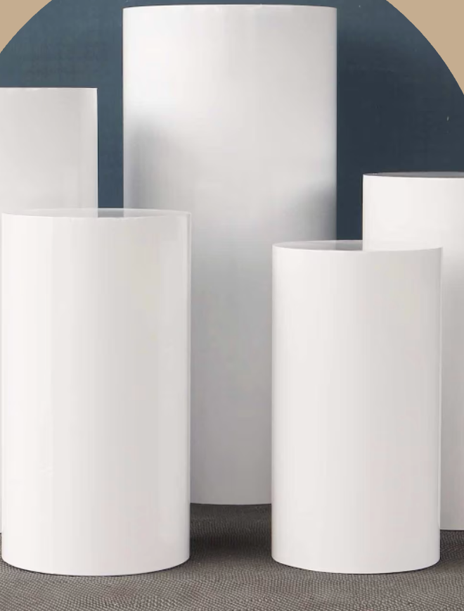 Five large white cylindrical display stands arranged on a gray surface against a dark wall, with uneven heights.