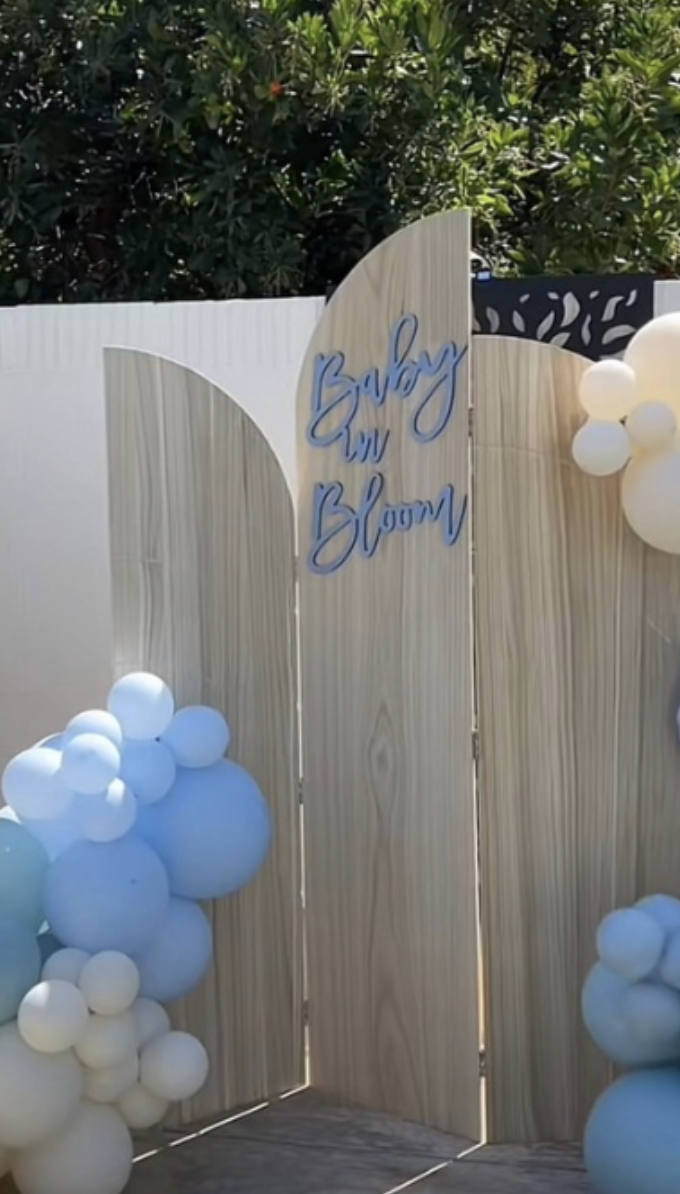 Decorative wooden panel backdrop with the phrase "Baby in Bloom" in blue cursive letters, surrounded by clusters of white and light blue balloons, set outdoors with green bushes in the background.